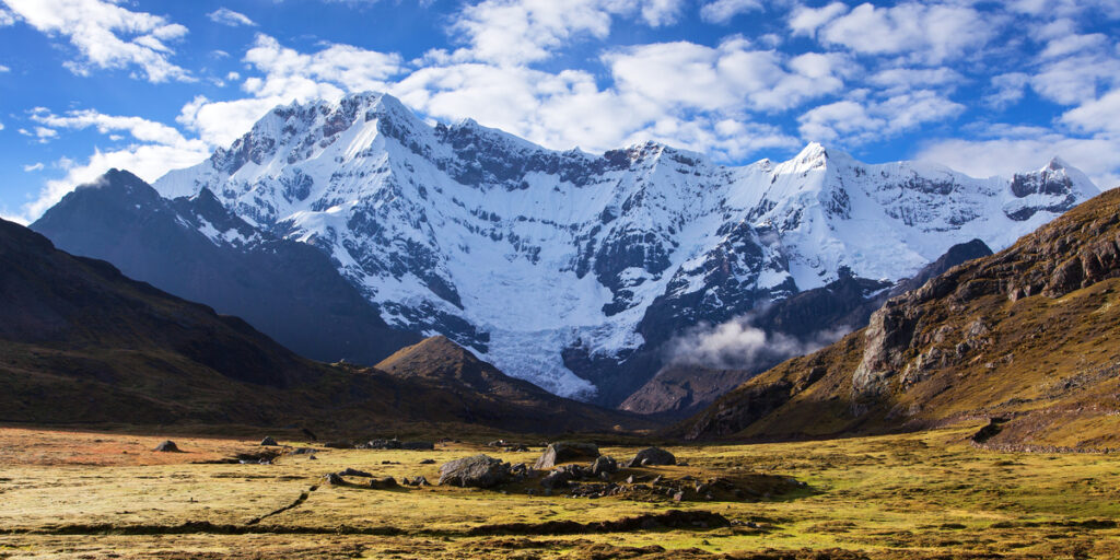 The snowcapped peak of Ausangate rises sharply beneath a bright blue sky with scattered clouds, surrounded by rocky terrain and high-altitude Andean plains.