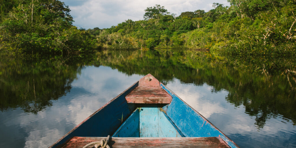 A view from a canoe floating down the wide Amazon River on a bright sunny day, surrounded by dense green rainforest trees lining both riverbanks under a clear blue sky.