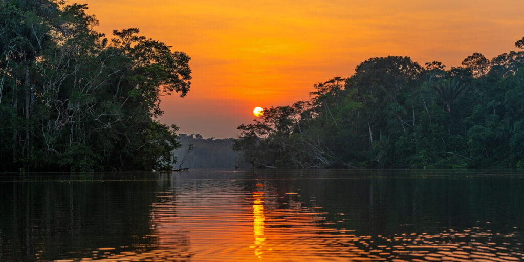 A wide view of the Amazon River at sunset, with the sky glowing deep orange. The river is calm and reflects the bright sun, surrounded by thick, green rainforest trees stretching along both banks.