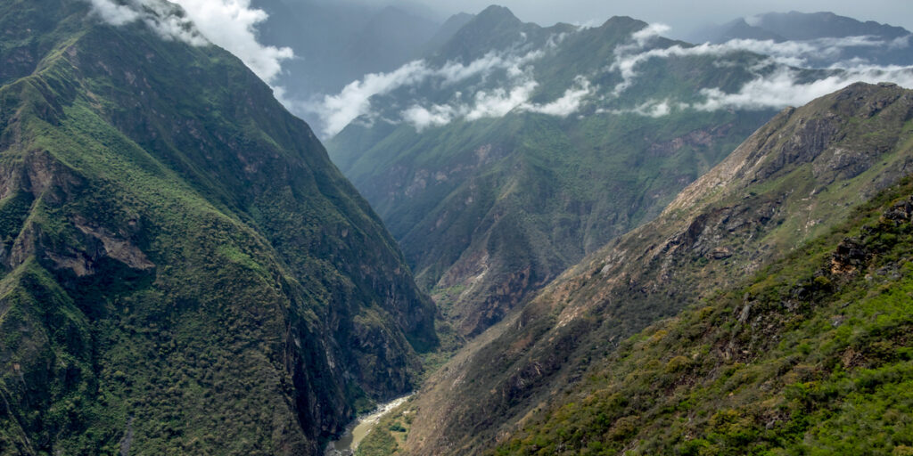A deep, green canyon with rugged slopes and misty clouds weaving through the peaks—Apurímac Canyon in Peru, with a winding river far below.

