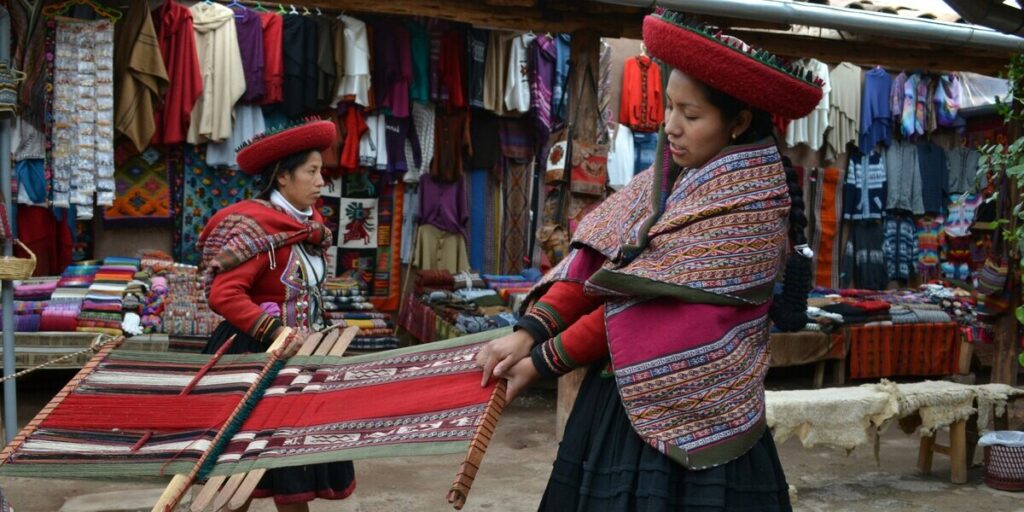 Two Andean women in traditional dress weave a vibrant textile by hand in an open-air Cusco market, surrounded by colorful fabrics, ponchos, and artisan crafts.