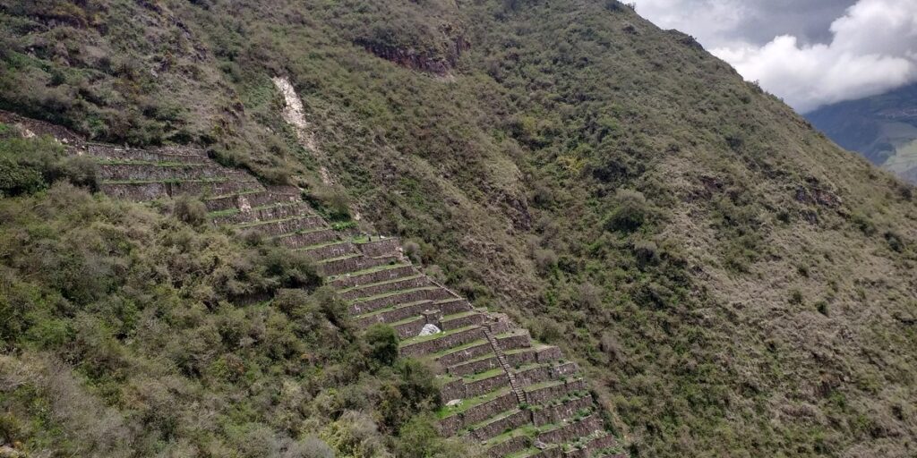 Ancient Inca agricultural terraces built into a steep, green mountainside at Choquequirao, partially hidden by dense vegetation and dramatic skies above.