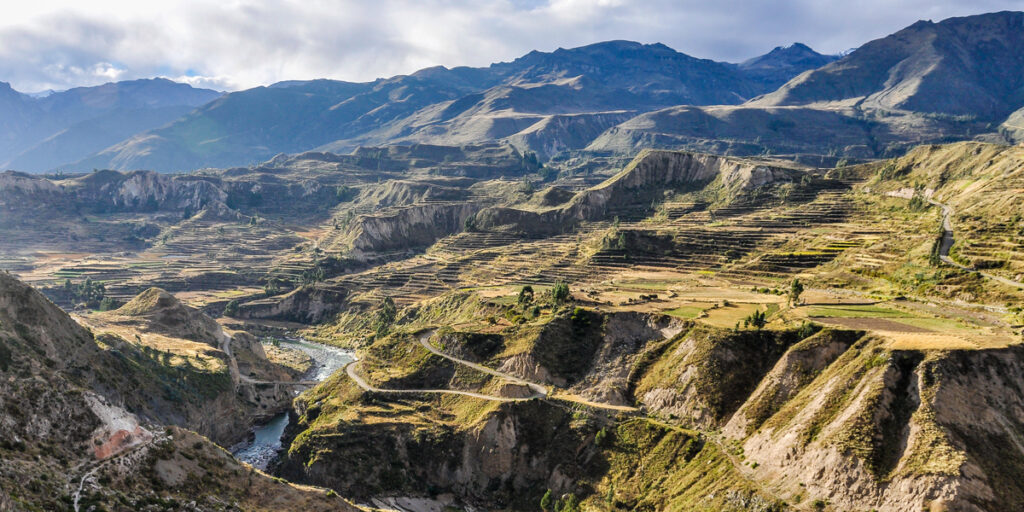 A deep canyon with steep rocky cliffs and green terraced slopes, with a winding river far below and distant mountains under a bright sky.