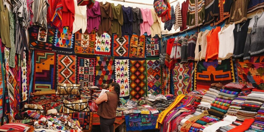 A local woman holding a baby stands inside a colorful market stall in Cusco, surrounded by vibrant textiles, alpaca sweaters, and traditional Andean weavings.