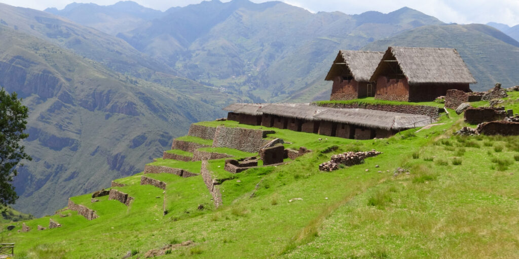 Ancient Inca ruins of Huchuy Qosqo nestled in the Andes mountains, showing thatched-roof stone structures, terraced agricultural fields, and panoramic views of the green valley below.