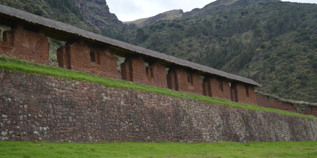 A long, ancient stone wall made of large, irregular rocks forms the base of several red-brown stone structures with small window-like openings, all topped with a dark, flat roof. Green grass slopes gently in front of the wall, and behind the structures, steep, verdant mountains rise under an overcast sky.