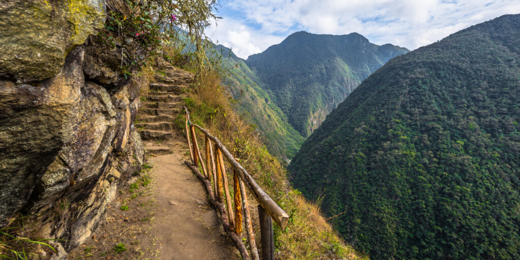 Stone steps and a dirt path bordered by a rustic wooden railing follow the edge of a cliff along the Inca Trail, with forested mountains rising in the background under a partly cloudy sky.
