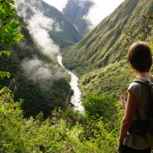A female hiker with a backpack stands on a trail surrounded by dense green vegetation, gazing down at a river winding through the valley below.