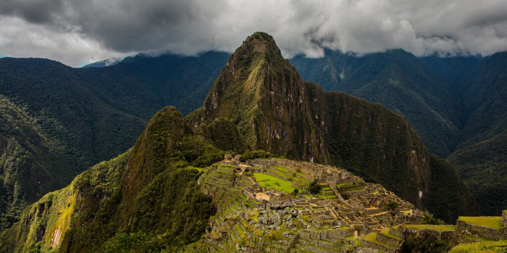 Stone terraces and ancient Inca buildings of Machu Picchu perched on a mountain ridge, surrounded by steep green peaks and misty clouds.