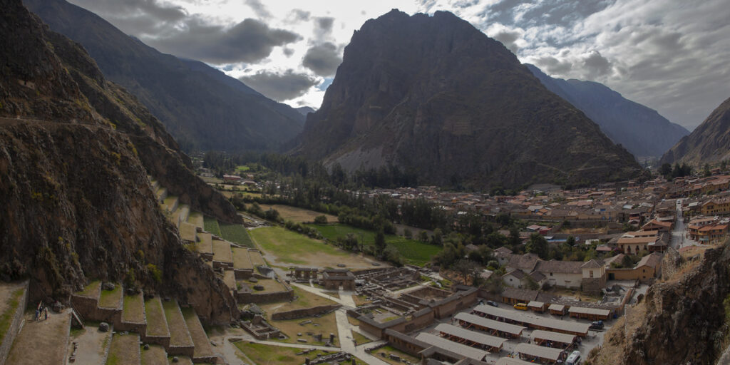 Panoramic view of Ollantaytambo archaeological site with its agricultural terraces, ancient ruins, and the surrounding town nestled in a valley beneath towering Andean mountains.