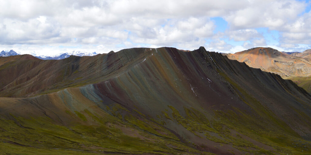 A panoramic view of Palccoyo Rainbow Mountain, showing several gently sloping hills striped with soft pastel colors like pink, yellow, and green, under a bright blue sky with scattered clouds.