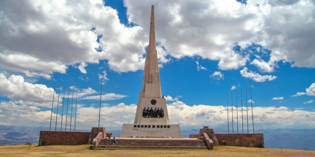 The Pampas de la Quinua Obelisk standing in an open, beige-toned Andean landscape with a few people nearby, dramatic clouds overhead, and distant hills surrounding the scene in Ayacucho.