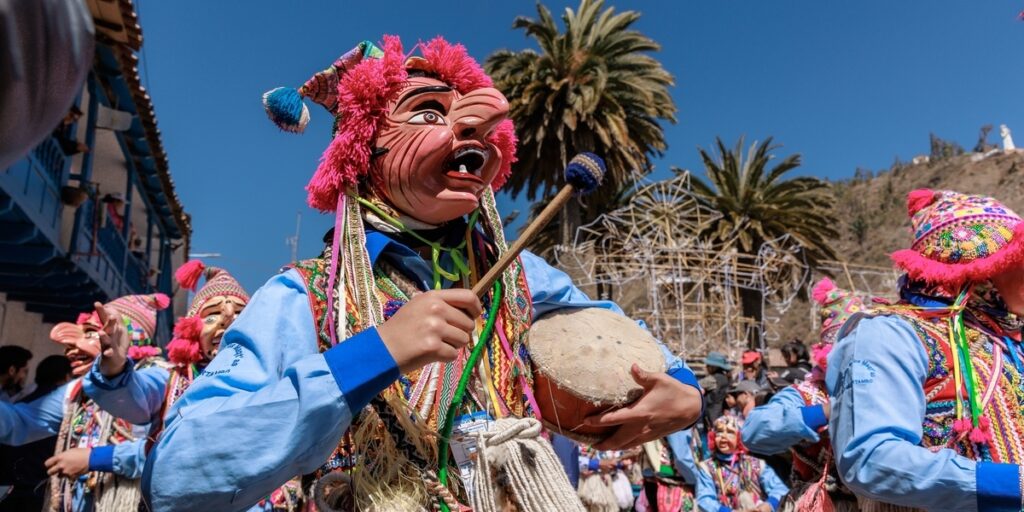 A festival performer in a colorful traditional costume stands in front of musicians playing wind instruments during the Virgen del Carmen celebration in Paucartambo, Peru.