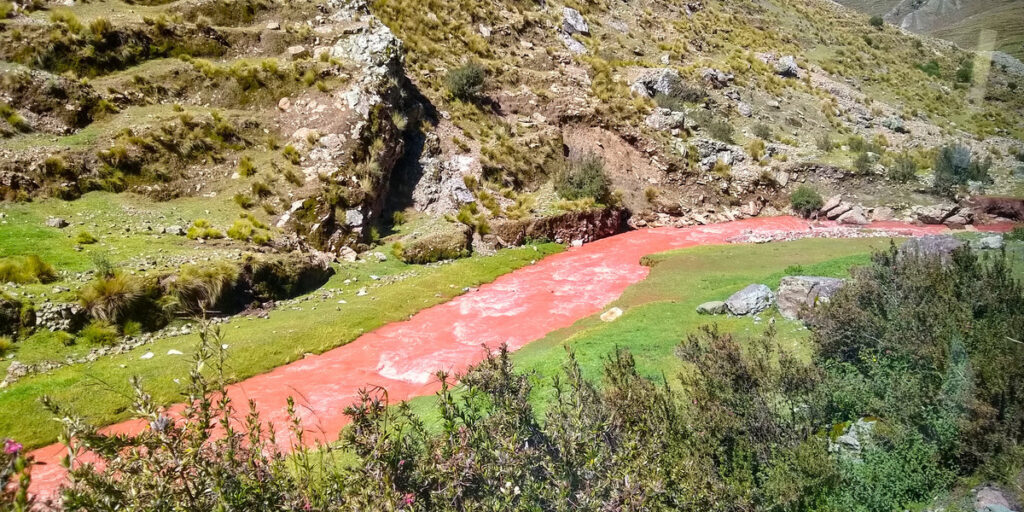A narrow red-colored river winds through a rocky Andean valley near Palccoyo, with rusty-red water contrasting against pale green grass and layered hills in the distance under a cloudy sky.