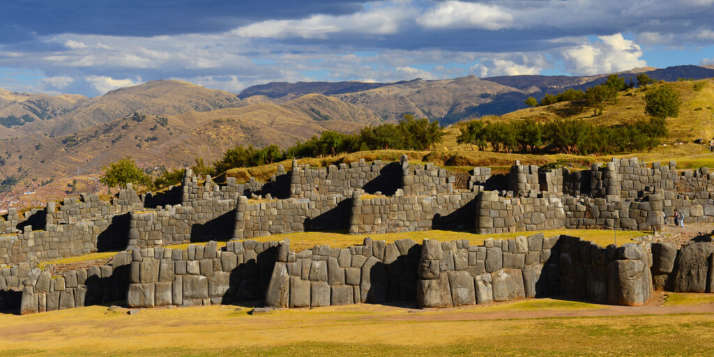 Massive zigzagging stone walls of Sacsayhuamán set against a grassy hilltop, with Cusco city and distant Andean mountains in the background.