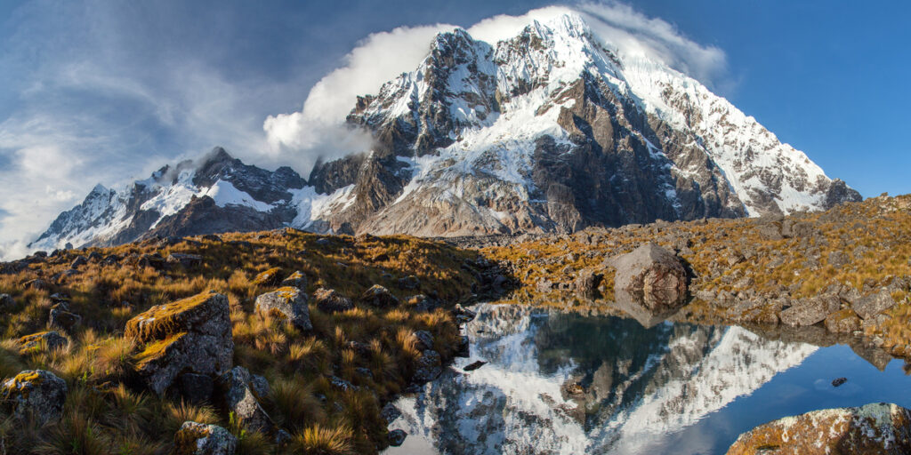A snow-covered Salkantay Mountain rises sharply above rugged, rocky terrain, with a dramatic contrast between white glaciers and the surrounding green valleys.