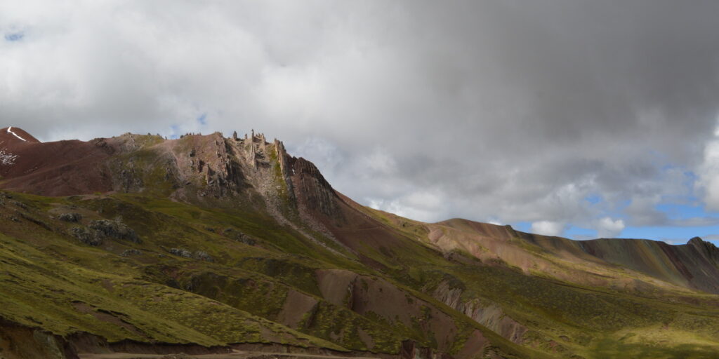 Jagged rock formations rise from a sloping green hillside under a blue sky, with snowcapped mountains in the distant background.