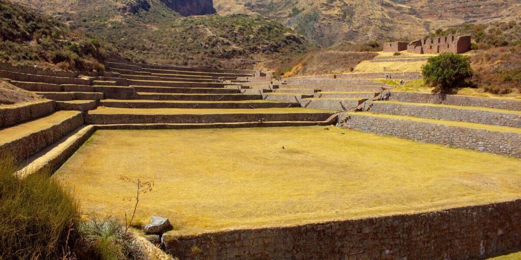 Wide view of the Inca terraces and stone walls of Tipón, an archaeological site in the Andes with dry yellowish grass and surrounding hills.