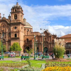 Cusco Cathedral towers over the vibrant Plaza de Armas, where locals and tourists relax on benches surrounded by gardens, colonial buildings, and mountain views.