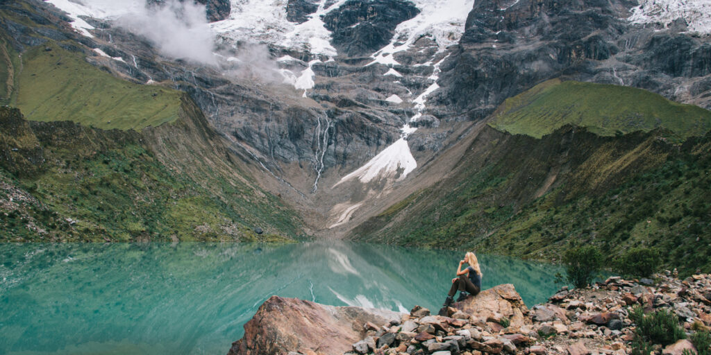 A young woman sits on a rock overlooking the turquoise waters of Lake Humantay, surrounded by towering mountains and vibrant wildflowers under a bright sky.