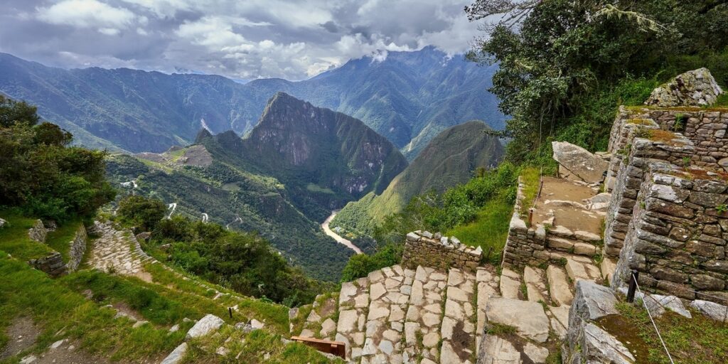 The ancient stone pathway of Intipunku (Sun Gate) overlooking the valley and Machu Picchu in the distance, surrounded by dramatic Andean mountains.