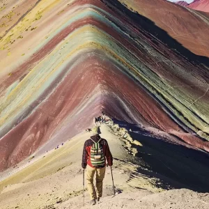 A hiker with trekking poles walks along a ridge towards the colorful, striped slopes of Rainbow Mountain (Vinicunca) in Peru.
