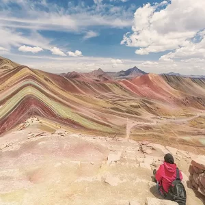 A person sits on a viewpoint overlooking the panoramic expanse of the colorful, striped mountains of Palccoyo, Peru.