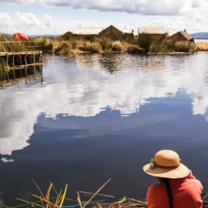 A person in a straw hat sits by a boat, looking out at the Uros floating islands and their reflection on Lake Titicaca.