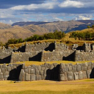 Massive zigzagging stone walls of Sacsayhuamán set against a grassy hilltop, with Cusco city and distant Andean mountains in the background.