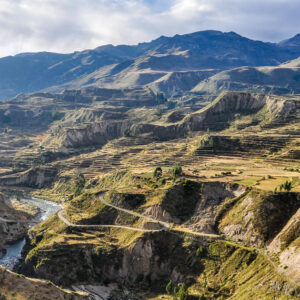 Panoramic view of the Colca Valley in southern Peru, showing terraced mountains, green fields, and the deep canyon under a bright sky.